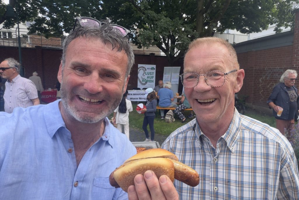 Ulrich Naust (rechts), Grillmeister in Betreuung bei BECKER Hörakustik (Filiale Puderbach), gemeinsam mit Dan Hilgert-Becker, Hör-Implantat-Spezialist von BECKER Hörakustik, an der Grillstation des CI-Sommerfestes 2026. Neben der kulinarischen Versorgung stand auch hier der persönliche Austausch im Mittelpunkt – zwischen Versorgungserfahrung, Alltagsperspektiven und langjähriger Verbundenheit zur CI-Community.
